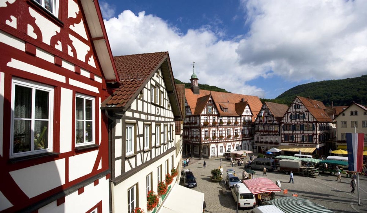 Blick auf den Marktplatz in Bad Urach mit malerischen Fachwerkhäusern und bunten Marktständen unter einem blauen Himmel mit Wolken., © Bad Urach Tourismus Blick auf den Marktplatz in Bad Urach mit malerischen Fachwerkhäusern und bunten Marktständen unter einem blauen Himmel mit Wolken., © Bad Urach Tourismus