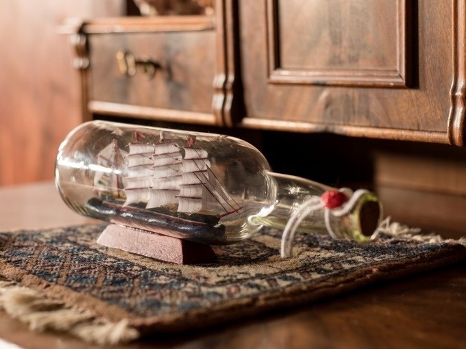 Ein Schiffsmodell in einer Flasche steht auf einem kleinen Teppich vor einem antiken Holzmöbel. Das Schiff hat weiße Segel., © ExitGames Stuttgart Ein Schiffsmodell in einer Flasche steht auf einem kleinen Teppich vor einem antiken Holzmöbel. Das Schiff hat weiße Segel., © ExitGames Stuttgart