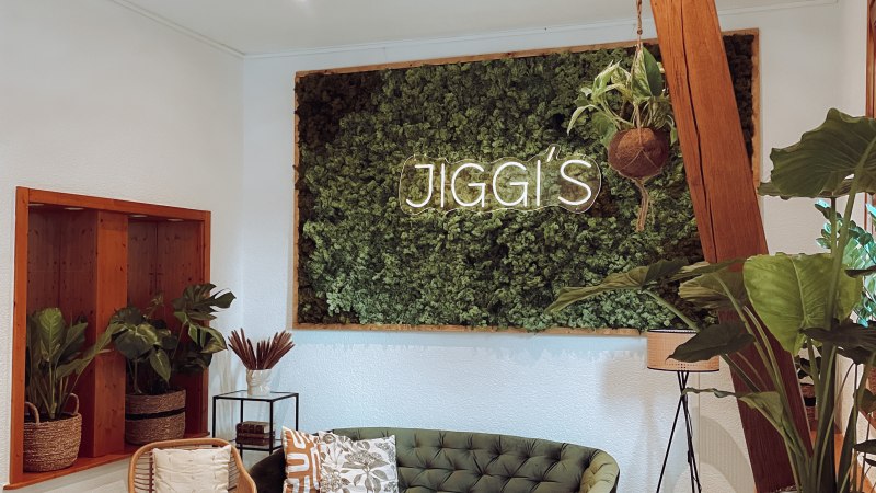 Gem&uuml;tlicher Raum mit gr&uuml;ner Couch, Rattanm&ouml;beln und Pflanzen. An der Wand ein Schild mit der Aufschrift 'Jiggi's'. Helle, einladende Atmosph&auml;re., &copy; Jiggis
