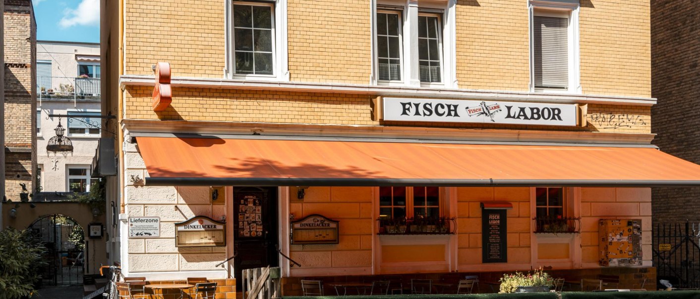 Ein Restaurant namens Fischlabor mit orangener Markise und Sitzgelegenheiten im Freien. Das Gebäude hat gelbe Ziegelwände und weiße Fensterrahmen., © SMG, Sarah Schmid