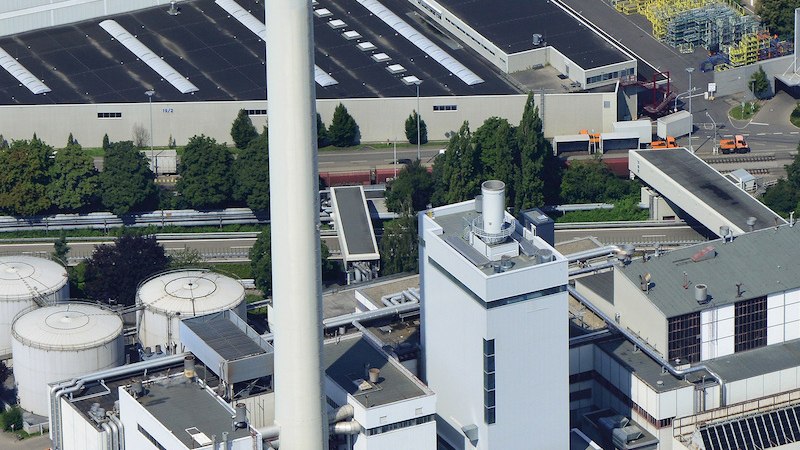 Luftaufnahme eines Industriekomplexes mit hohem Schornstein, mehreren Gebäuden und Tanks. Grünflächen und Straßen umgeben die Anlage., © Mercedes-Benz AG Luftaufnahme eines Industriekomplexes mit hohem Schornstein, mehreren Gebäuden und Tanks. Grünflächen und Straßen umgeben die Anlage., © Mercedes-Benz AG
