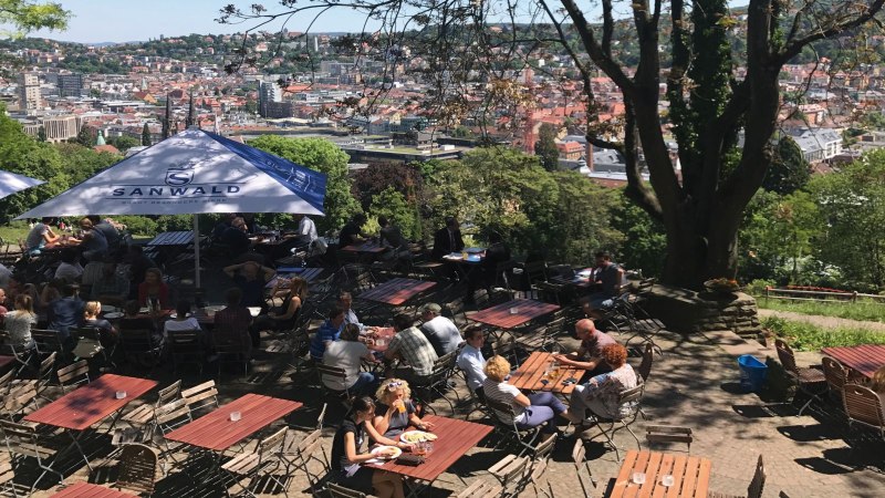 Ein Biergarten auf der Karlsh&ouml;he mit Blick auf Stuttgart. Menschen sitzen an Tischen unter Sonnenschirmen, umgeben von B&auml;umen und einer Stadtansicht., &copy; Stuttgart-Marketing GmbH