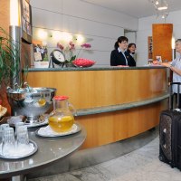 Eine Hotelrezeption mit zwei Mitarbeiterinnen, die einer &auml;lteren Frau mit Koffer helfen. Ein Tisch mit &Auml;pfeln und Getr&auml;nken steht im Vordergrund., &copy; Hotel Unger