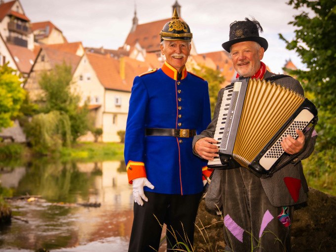 Zwei Männer in historischen Kostümen posieren vor einem malerischen Fluss. Einer trägt eine blaue Uniform, der andere spielt Akkordeon. Im Hintergrund sind Fachwerkhäuser zu sehen., © Stadt Besigheim Zwei Männer in historischen Kostümen posieren vor einem malerischen Fluss. Einer trägt eine blaue Uniform, der andere spielt Akkordeon. Im Hintergrund sind Fachwerkhäuser zu sehen., © Stadt Besigheim