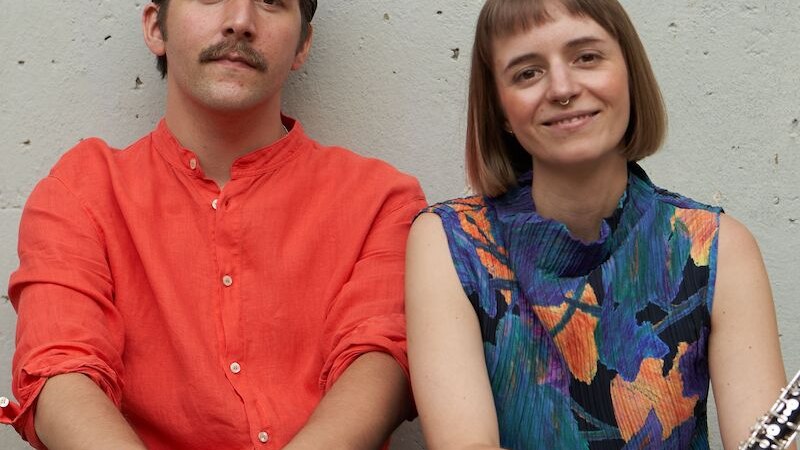 Zwei Personen sitzen vor einer Wand. Eine Person trägt ein rotes Hemd und hält Schlagzeugstöcke, die andere ein buntes Kleid und ein Musikinstrument., © Mauerwerk GmbH Zwei Personen sitzen vor einer Wand. Eine Person trägt ein rotes Hemd und hält Schlagzeugstöcke, die andere ein buntes Kleid und ein Musikinstrument., © Mauerwerk GmbH