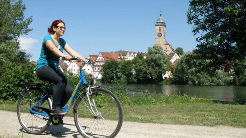Eine Frau fährt Fahrrad am Neckar in Nürtingen. Im Hintergrund sind eine Kirche und Fachwerkhäuser zu sehen., © Stadt Nürtingen