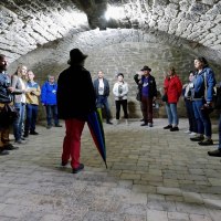 Menschen stehen in einem Gew&ouml;lbekeller aus Stein, h&ouml;ren einer Person mit Hut zu. Der Raum ist mit Kopfsteinpflaster ausgelegt., &copy; Clemens Scholl