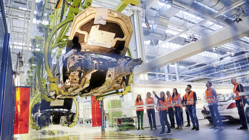 Besucher in orangefarbenen Westen beobachten die Fahrzeugmontage in einer Mercedes-Benz Produktionshalle in Sindelfingen., © Mercedes-Benz AG