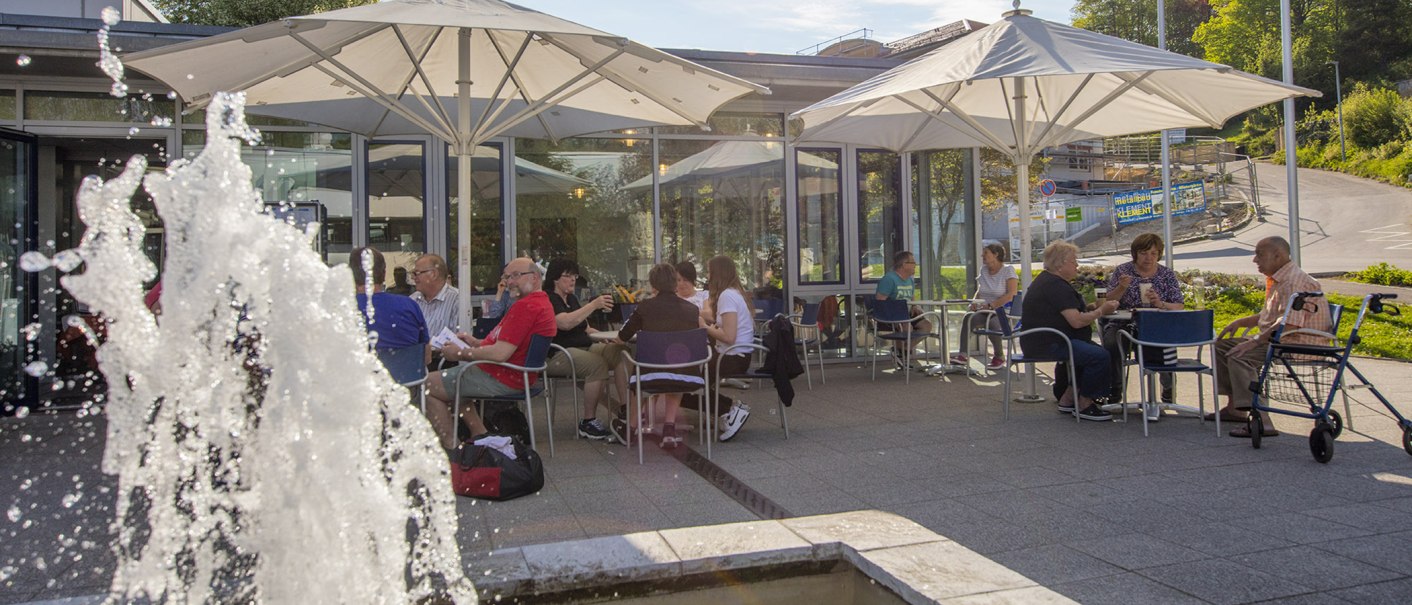 Menschen genießen das Café Vinzenz Therme im Freien, sitzen unter Sonnenschirmen an Tischen. Ein Springbrunnen im Vordergrund sprüht Wasser., © Vinzenz Therme Bad Ditzenbach / Peter Flämig Menschen genießen das Café Vinzenz Therme im Freien, sitzen unter Sonnenschirmen an Tischen. Ein Springbrunnen im Vordergrund sprüht Wasser., © Vinzenz Therme Bad Ditzenbach / Peter Flämig