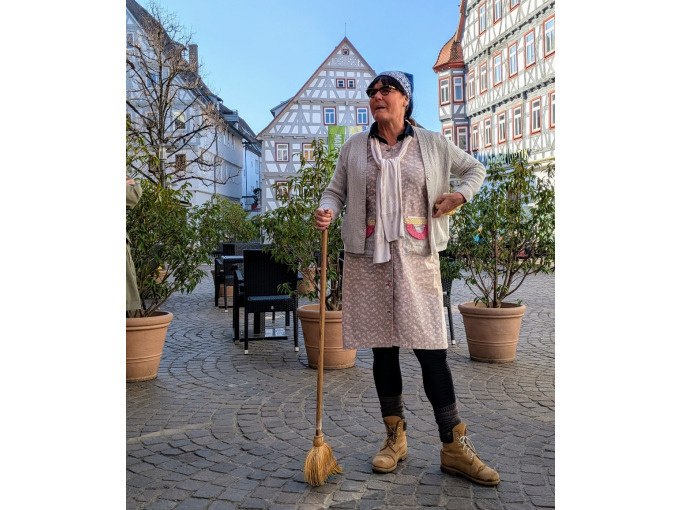 Person in traditioneller Kleidung mit Kopftuch und Besen steht auf einem gepflasterten Platz vor Fachwerkh&auml;usern., &copy; WTM GmbH Waiblingen
