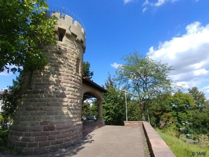 Ein steinerner Turm mit Zinnen steht neben einem Weg, umgeben von Bäumen unter blauem Himmel., © Stuttgart Marketing GmbH Ein steinerner Turm mit Zinnen steht neben einem Weg, umgeben von Bäumen unter blauem Himmel., © Stuttgart Marketing GmbH