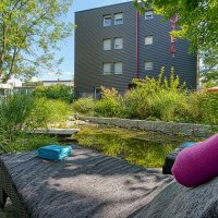 Ein moderner Garten mit Teich, umgeben von Pflanzen. Im Vordergrund ein Liegestuhl mit Handtüchern, im Hintergrund ein mehrstöckiges Gebäude., © Centro Hotels Ein moderner Garten mit Teich, umgeben von Pflanzen. Im Vordergrund ein Liegestuhl mit Handtüchern, im Hintergrund ein mehrstöckiges Gebäude., © Centro Hotels