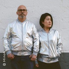 Zwei Personen in silbernen Jacken stehen vor einer wei&szlig;en Wand und blicken in verschiedene Richtungen., &copy; links im Bild