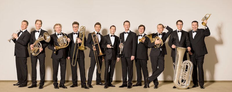Elf Männer in schwarzen Anzügen halten Blasinstrumente und posieren lächelnd vor einer neutralen Wand., © Schönblick gemeinnützige GmbH Elf Männer in schwarzen Anzügen halten Blasinstrumente und posieren lächelnd vor einer neutralen Wand., © Schönblick gemeinnützige GmbH
