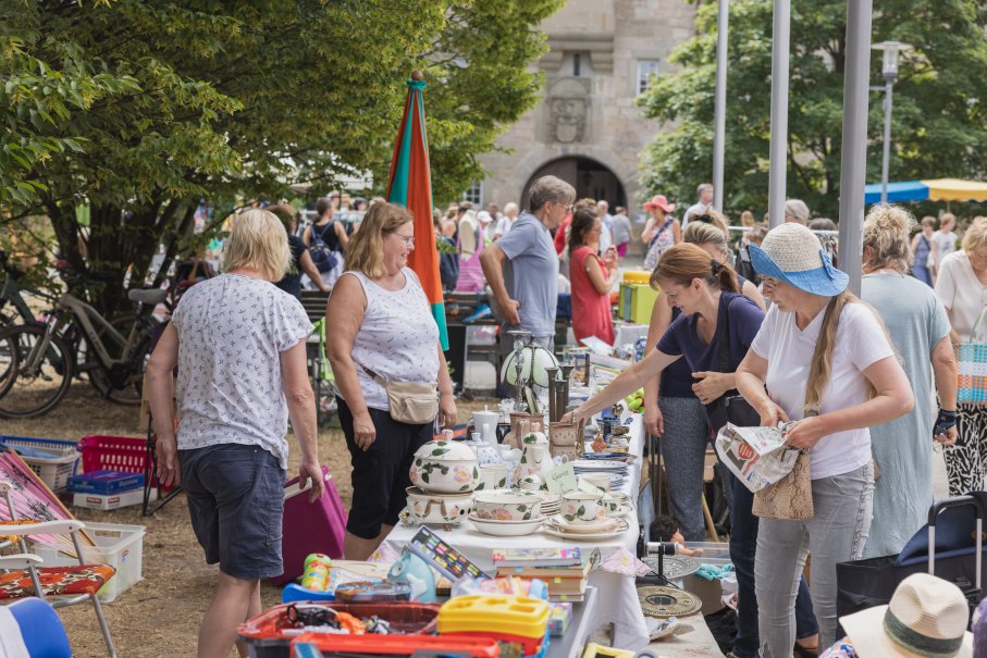 Flohmarktbesucher st&ouml;bern an einem Stand mit Geschirr und Spielzeug. Im Hintergrund sind weitere Menschen und B&auml;ume zu sehen., &copy; BEBOP MEDIA