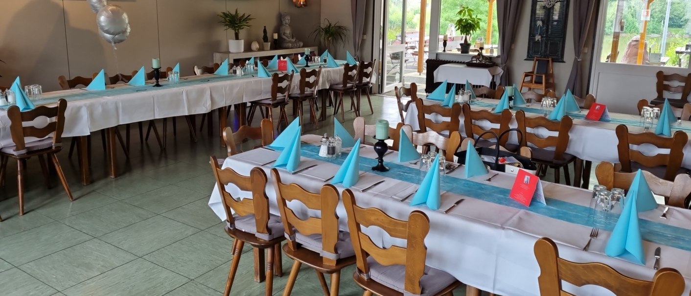 Festlich gedeckter Raum mit langen Tischen, blauen Servietten und Dekorationen. Große Fenster bieten Blick nach draußen. Luftballons und Pflanzen schmücken den Raum., © Gaststätte Goldbachsee Festlich gedeckter Raum mit langen Tischen, blauen Servietten und Dekorationen. Große Fenster bieten Blick nach draußen. Luftballons und Pflanzen schmücken den Raum., © Gaststätte Goldbachsee