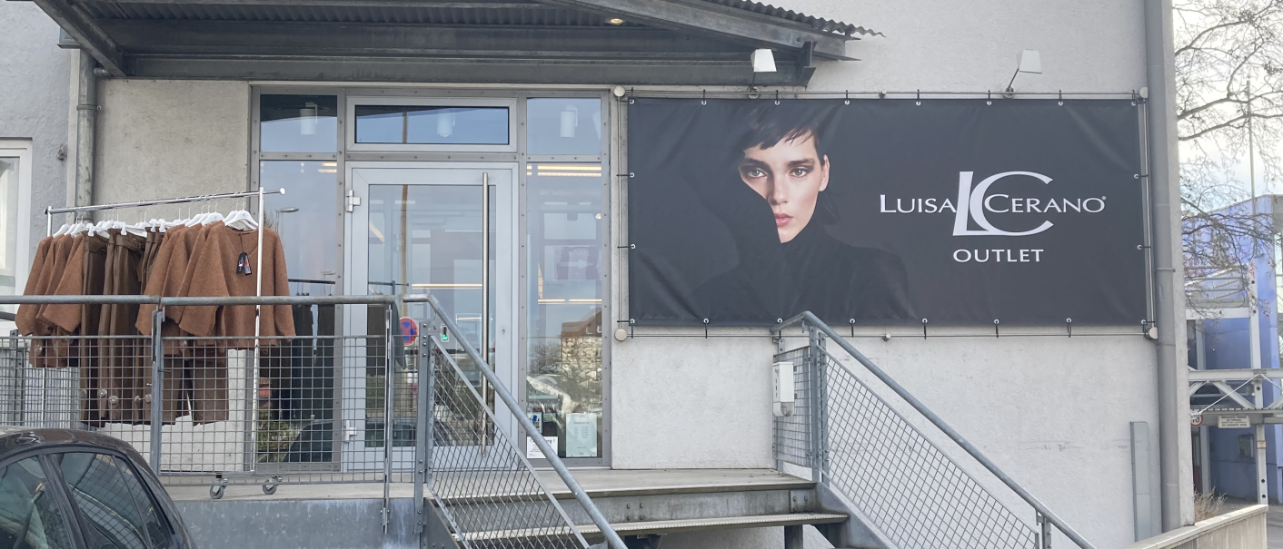 Eingang eines Luisa Cerano Outlets mit Treppe, Werbebanner und Kleidung auf einem Ständer. Ein Auto parkt daneben, Büsche im Vordergrund., © L. Besmer Eingang eines Luisa Cerano Outlets mit Treppe, Werbebanner und Kleidung auf einem Ständer. Ein Auto parkt daneben, Büsche im Vordergrund., © L. Besmer