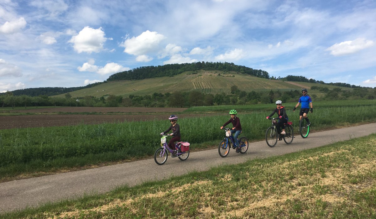 Eine Familie fährt auf einem Radweg durch eine ländliche Landschaft. Im Hintergrund ist ein bewachsener Hügel unter blauem Himmel zu sehen., © Land der 1000 Hügel - Kraichgau-Stromberg