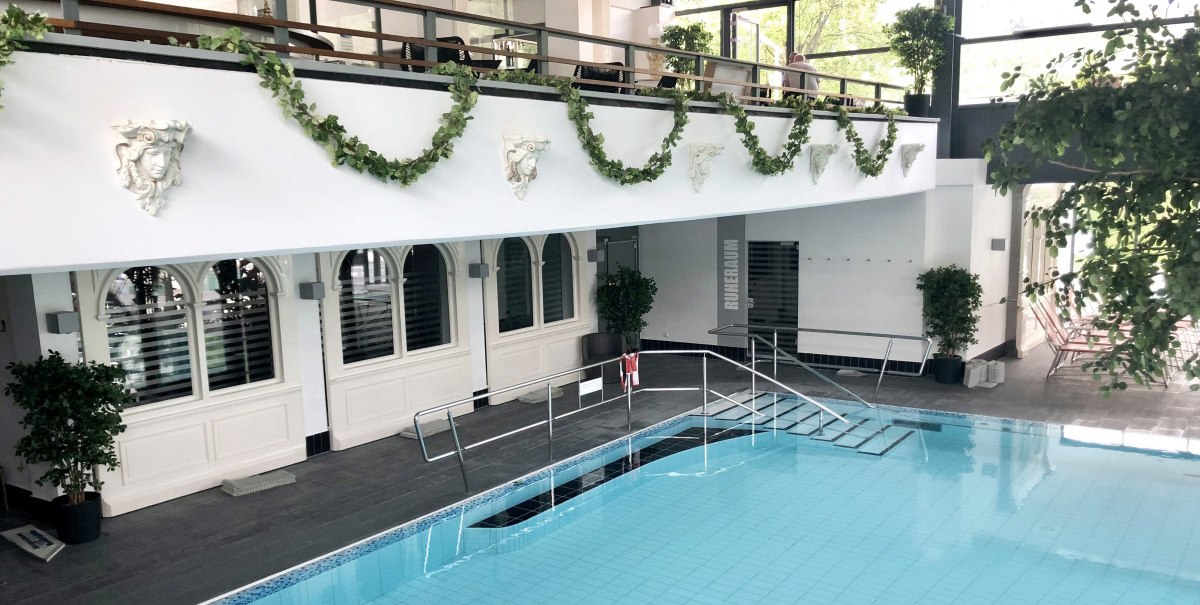 Elegantes Schwimmbad mit blauen Fliesen, Pflanzen und dekorativen Elementen an den Wänden. Obergeschoss mit Geländer und Kronleuchter., © AlbThermen Bad Urach