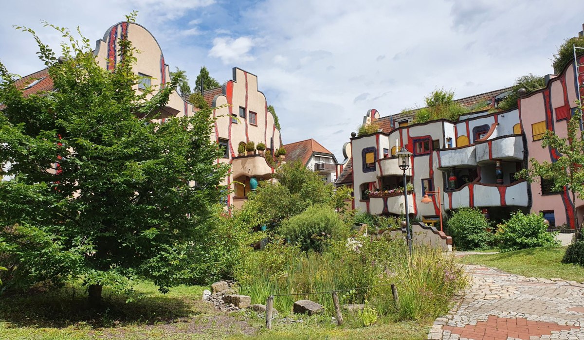 Buntes Gebäude im Hundertwasser-Stil mit geschwungenen Linien, Balkonen und viel Grün im Vordergrund. Ein gepflasterter Weg führt durch den Garten.