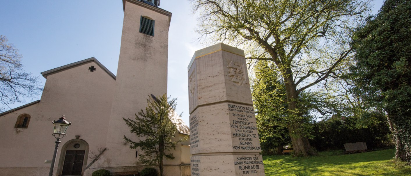 Die Evangelische Stiftskirche Bad Boll mit einem Denkmal im Vordergrund, umgeben von Bäumen und einer Laterne., © SMG, Achim Mende