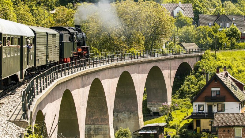 Eine Dampflokomotive zieht einen Zug über eine Brücke in einer grünen, ländlichen Landschaft. Menschen schauen aus den Fenstern und fotografieren., © SMG, Sarah Schmid Eine Dampflokomotive zieht einen Zug über eine Brücke in einer grünen, ländlichen Landschaft. Menschen schauen aus den Fenstern und fotografieren., © SMG, Sarah Schmid