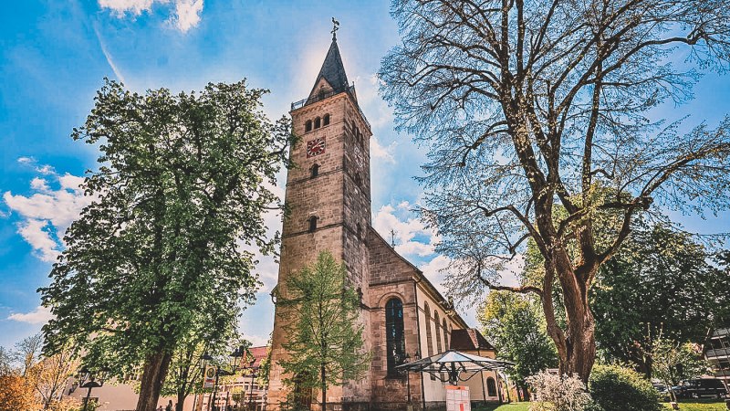 Eine Kirche mit hohem Turm, umgeben von Bäumen, bei sonnigem Wetter. Der Turm hat eine Uhr und ein spitzes Dach., © Stadt Welzheim