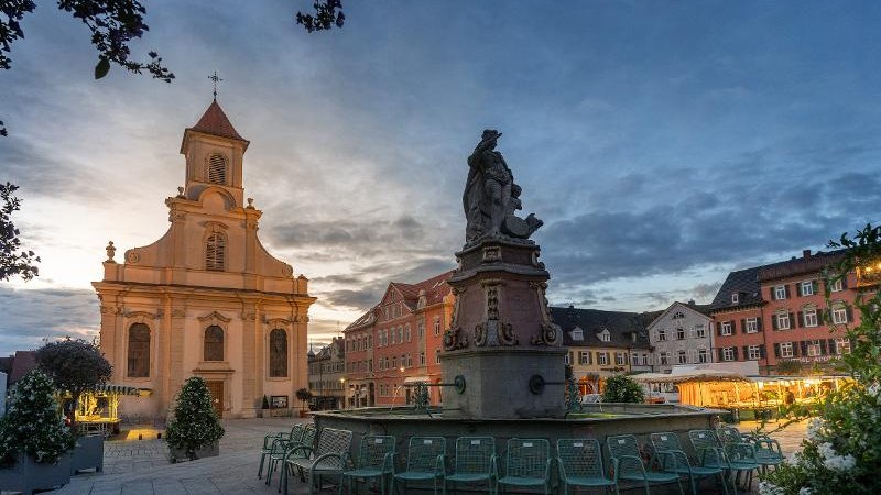 Abendstimmung auf einem Platz in Ludwigsburg. Eine Kirche und ein Brunnen sind im Vordergrund, umgeben von historischen Gebäuden und Stühlen., © © Martina Denker