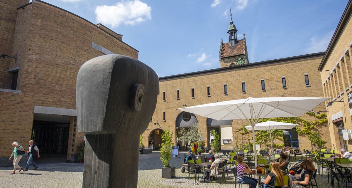 Ein Innenhof mit einer Holzskulptur im Vordergrund, Sitzbereich mit Sonnenschirmen und der Turm der Lutherkirche im Hintergrund., © Achim Mende Ein Innenhof mit einer Holzskulptur im Vordergrund, Sitzbereich mit Sonnenschirmen und der Turm der Lutherkirche im Hintergrund., © Achim Mende