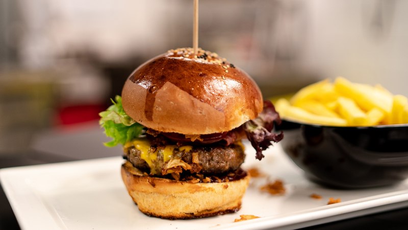 Ein saftiger Burger mit Salat und Käse auf einem Teller, daneben eine Schale mit Pommes frites., © Sensapolis GmbH