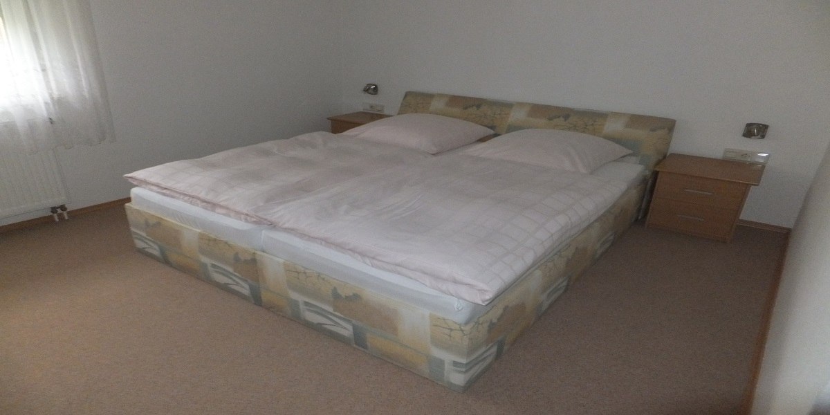 Ein einfach eingerichtetes Schlafzimmer mit einem Doppelbett, zwei Nachttischen und einem Fenster mit Vorhang., © Hotel Traube