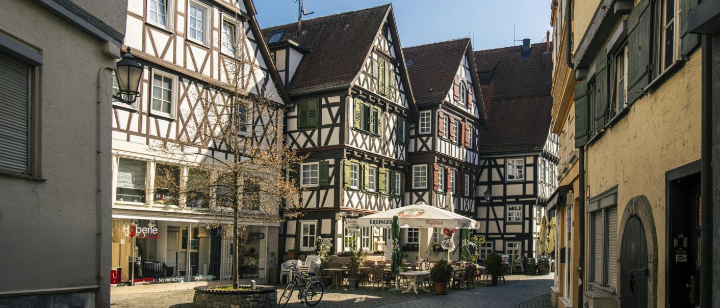 Fachwerkhäuser am Schorndorfer Marktplatz. Ein Café mit Sonnenschirmen und Tischen steht im Vordergrund. Die Szene wirkt einladend und historisch., © SMG, Sarah Schmid Fachwerkhäuser am Schorndorfer Marktplatz. Ein Café mit Sonnenschirmen und Tischen steht im Vordergrund. Die Szene wirkt einladend und historisch., © SMG, Sarah Schmid