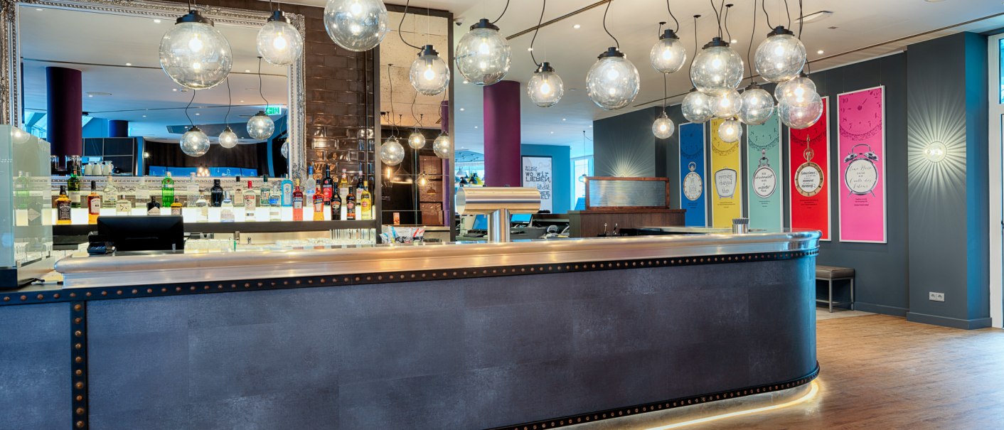 Moderne Bar mit hängenden Glaskugellampen, Spiegel und bunter Wanddekoration. Flaschen und Gläser stehen auf der Theke., © Premier Inn Germany