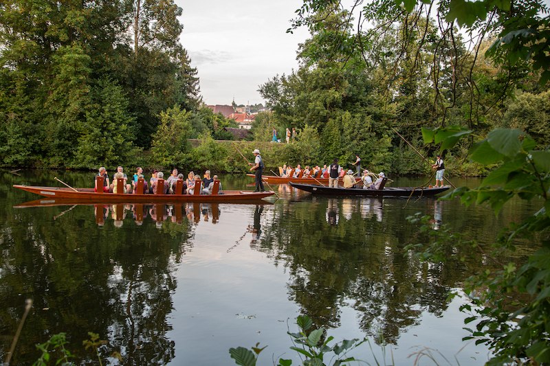 Zwei Boote mit Musikern fahren auf einem ruhigen Fluss, umgeben von üppigem Grün. Die Szene wirkt friedlich und idyllisch., © Stadtverwaltung Vaihingen an der Enz Zwei Boote mit Musikern fahren auf einem ruhigen Fluss, umgeben von üppigem Grün. Die Szene wirkt friedlich und idyllisch., © Stadtverwaltung Vaihingen an der Enz