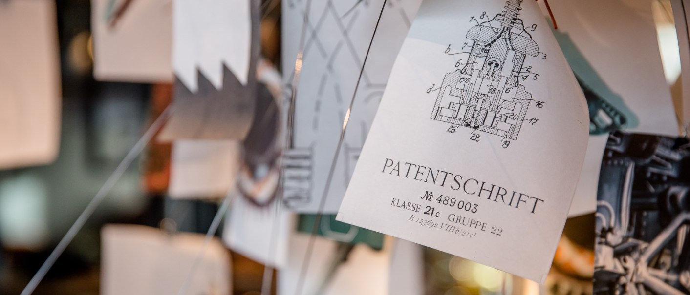 Nahaufnahme einer Patentschrift mit Zeichnung, umgeben von weiteren Papieren und Zeichnungen., © TOMAS