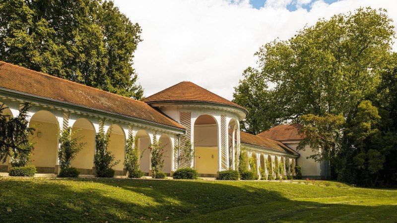 Die Wandelhalle in Bad Boll zeigt elegante Arkaden und ein rotes Ziegeldach, eingebettet in eine grüne Landschaft mit Bäumen und Rasen., © SMG, Sarah Schmid Die Wandelhalle in Bad Boll zeigt elegante Arkaden und ein rotes Ziegeldach, eingebettet in eine grüne Landschaft mit Bäumen und Rasen., © SMG, Sarah Schmid