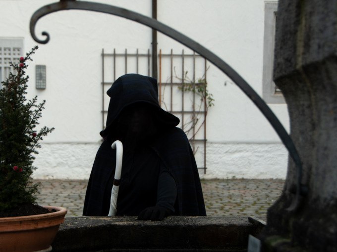 Vermummte Gestalt mit Kapuze und weißem Stock vor einem Brunnen in einem gepflasterten Hof., © Stadt Waldenbuch Vermummte Gestalt mit Kapuze und weißem Stock vor einem Brunnen in einem gepflasterten Hof., © Stadt Waldenbuch