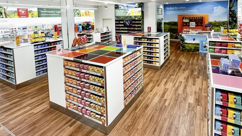 Ein heller Schokoshop mit bunten Ritter Sport Tafeln in Regalen, Holzboden und Werbepostern an den Wänden., © Alfred Ritter GmbH & Co. KG