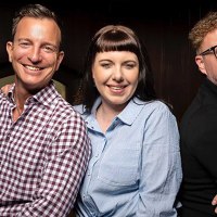 Drei Personen stehen l&auml;chelnd nebeneinander vor einer Holzwand. Sie tragen Freizeitkleidung und wirken fr&ouml;hlich., &copy; Zumbecher