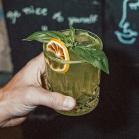 Eine t&auml;towierte Hand h&auml;lt ein gr&uuml;nes Getr&auml;nk mit Basilikum und Orangenscheibe. Im Hintergrund ist eine Tafel mit Zeichnungen und Schrift zu sehen., &copy; Bruno, Stuttgart
