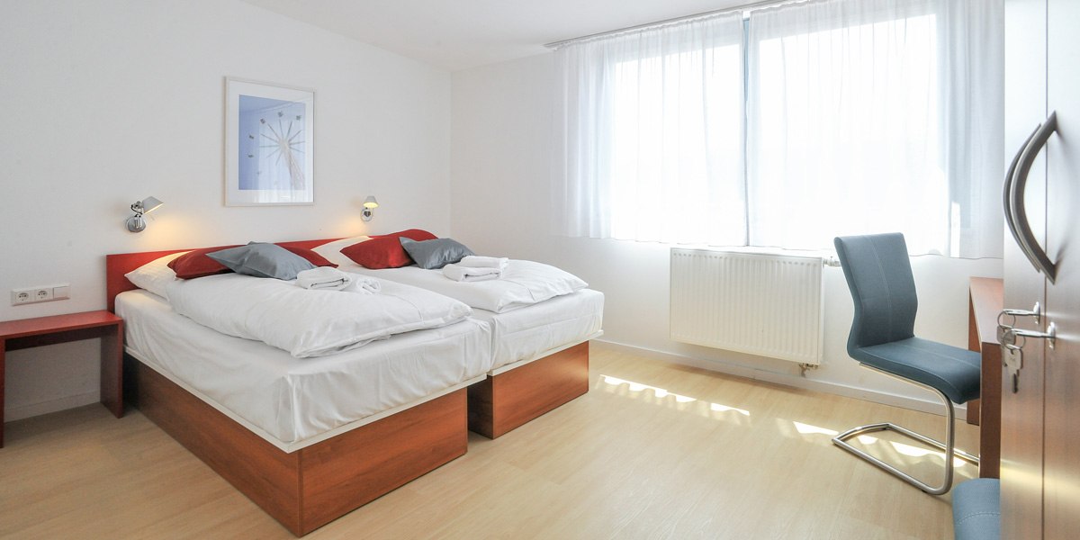 Helles Doppelzimmer mit zwei Einzelbetten, roten und grauen Kissen, einem Schreibtisch mit Stuhl und einem großen Fenster mit weißen Vorhängen., © Gästehaus ABZ Stuttgart Helles Doppelzimmer mit zwei Einzelbetten, roten und grauen Kissen, einem Schreibtisch mit Stuhl und einem großen Fenster mit weißen Vorhängen., © Gästehaus ABZ Stuttgart