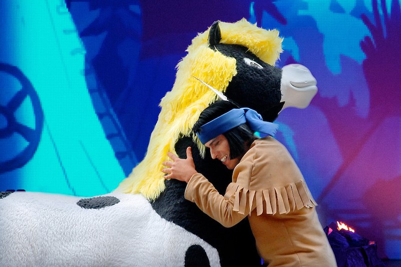 Ein Schauspieler in einem Yakari-Kostüm umarmt ein großes Plüschpferd. Der Hintergrund ist bunt und zeigt eine animierte Szene., © Whynot Events UG Ein Schauspieler in einem Yakari-Kostüm umarmt ein großes Plüschpferd. Der Hintergrund ist bunt und zeigt eine animierte Szene., © Whynot Events UG