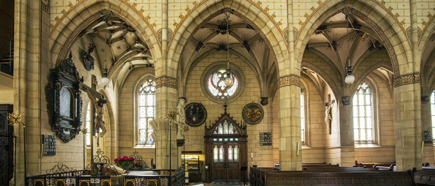 Innenansicht der Amanduskirche in Bad Urach mit gotischen B&ouml;gen, Kirchenb&auml;nken und einem Kruzifix. Buntglasfenster und dekorative Elemente sind sichtbar., &copy; Stuttgart-Marketing GmbH, Sarah Schmid