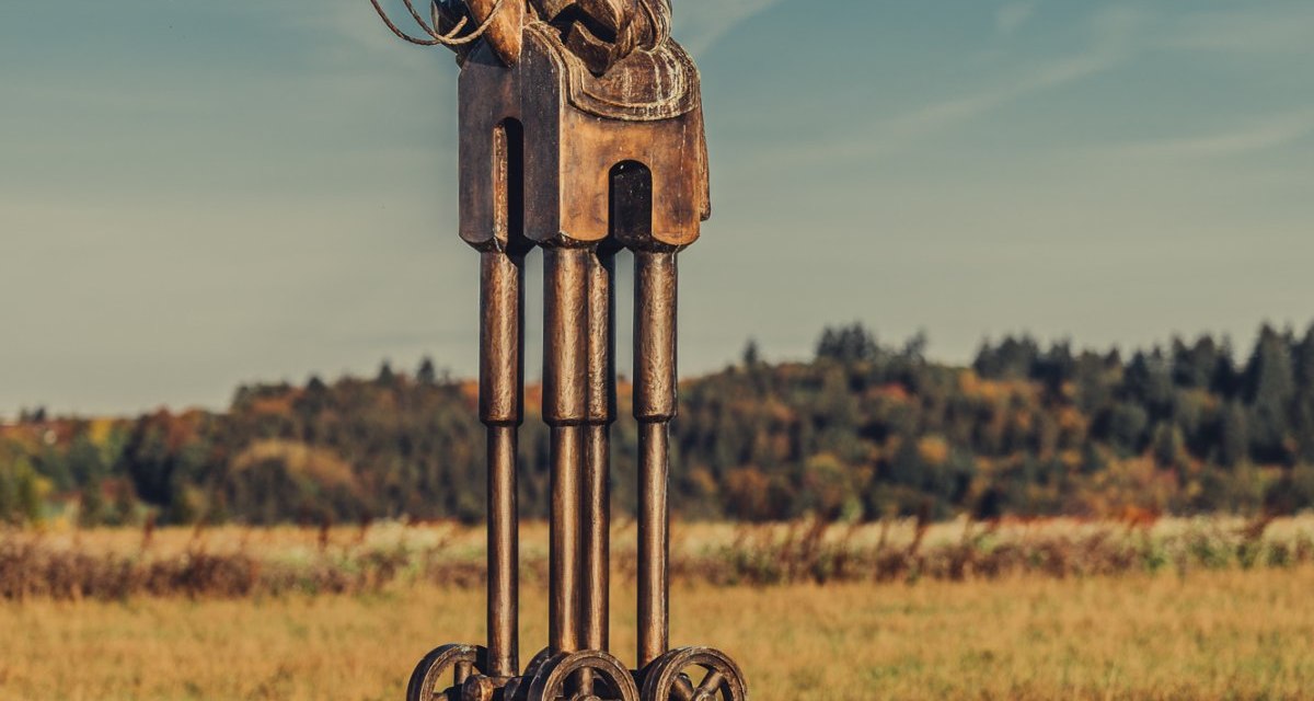 Metallskulptur eines Reiters auf einem Pferd mit Rädern, auf einem Sockel in einer Wiese unter blauem Himmel., © Landratsamt Ostalbkreis Metallskulptur eines Reiters auf einem Pferd mit Rädern, auf einem Sockel in einer Wiese unter blauem Himmel., © Landratsamt Ostalbkreis