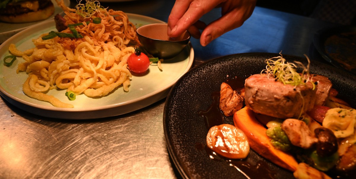 Zwei Teller mit kunstvoll angerichteten Speisen, darunter Sp&auml;tzle und Fleisch. Eine Hand richtet die Gerichte auf einem Tisch an.