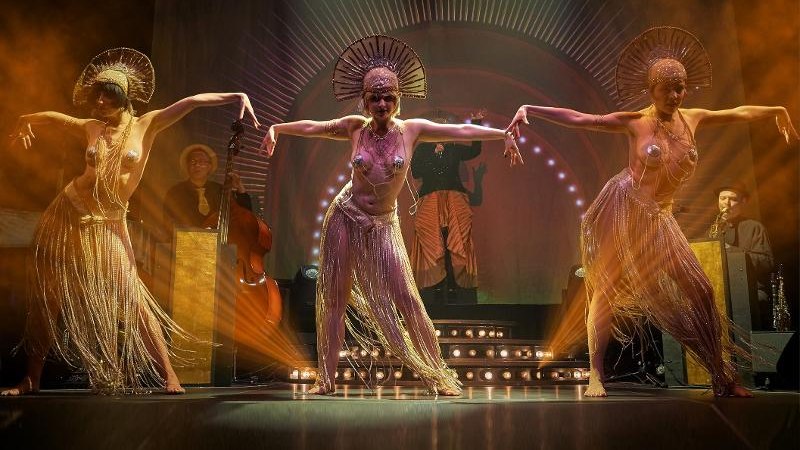 Drei Tänzerinnen in glitzernden Kostümen tanzen auf einer Bühne. Im Hintergrund sind Musiker und eine beleuchtete Kulisse zu sehen., © Backnang Drei Tänzerinnen in glitzernden Kostümen tanzen auf einer Bühne. Im Hintergrund sind Musiker und eine beleuchtete Kulisse zu sehen., © Backnang