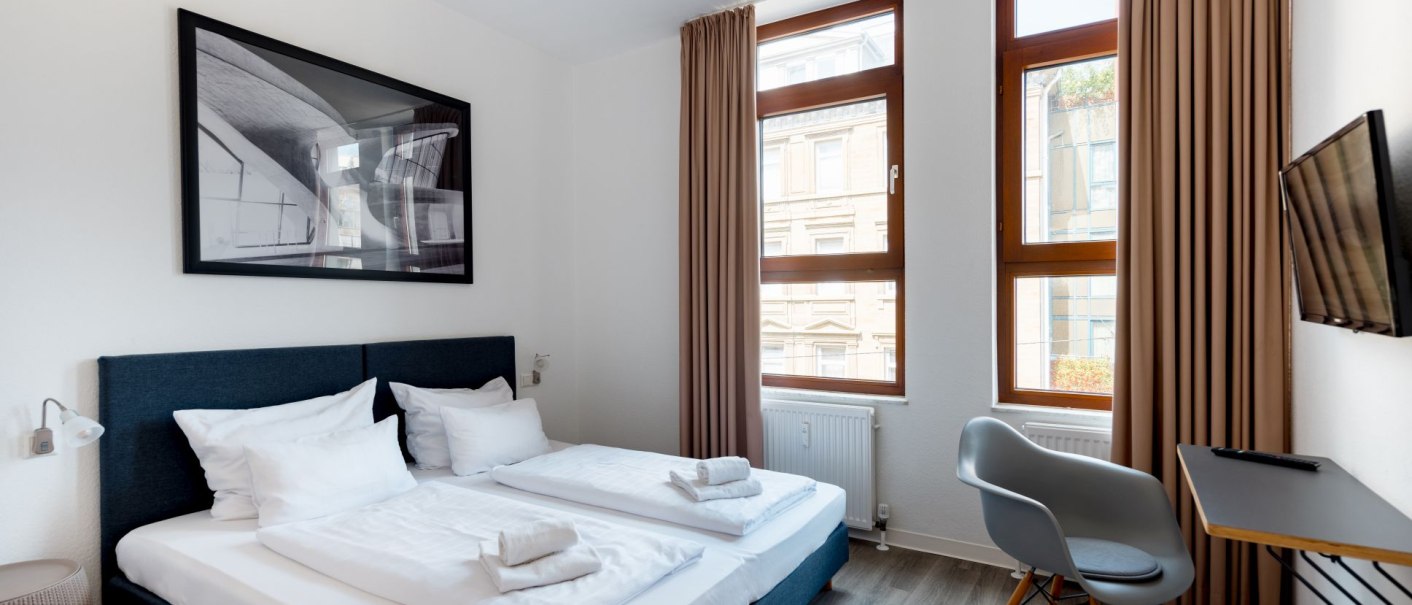 Modernes Doppelzimmer mit zwei Betten, Bild an der Wand, kleinem Tisch und Stuhl. Große Fenster mit Vorhängen., © Hotel Olgaeck