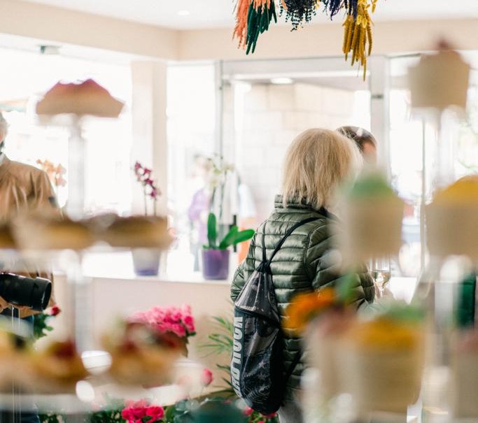 Ein Caf&eacute; mit h&auml;ngenden Trockenblumen und unscharfen Desserts im Vordergrund. Menschen im Hintergrund, umgeben von Pflanzen und Blumen., &copy; cMagnolia