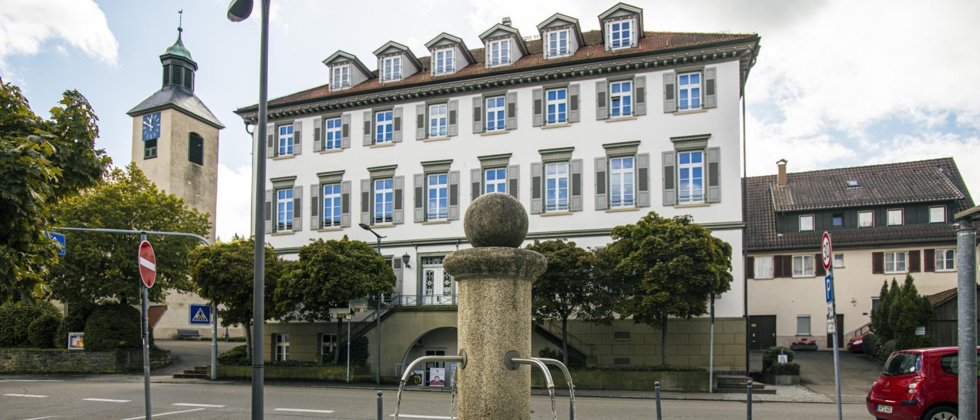 Das Rathaus von Bad Boll mit einem Brunnen im Vordergrund und einem Kirchturm im Hintergrund bei bewölktem Himmel., © Stuttgart-Marketing GmbH, Sarah Schmid