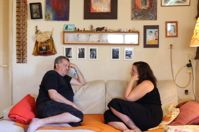 Zwei Personen sitzen auf einem Sofa in einem gemütlichen Wohnzimmer. An der Wand hängen Bilder und Fotos. Sie unterhalten sich entspannt., © Kunsthalle Göppingen